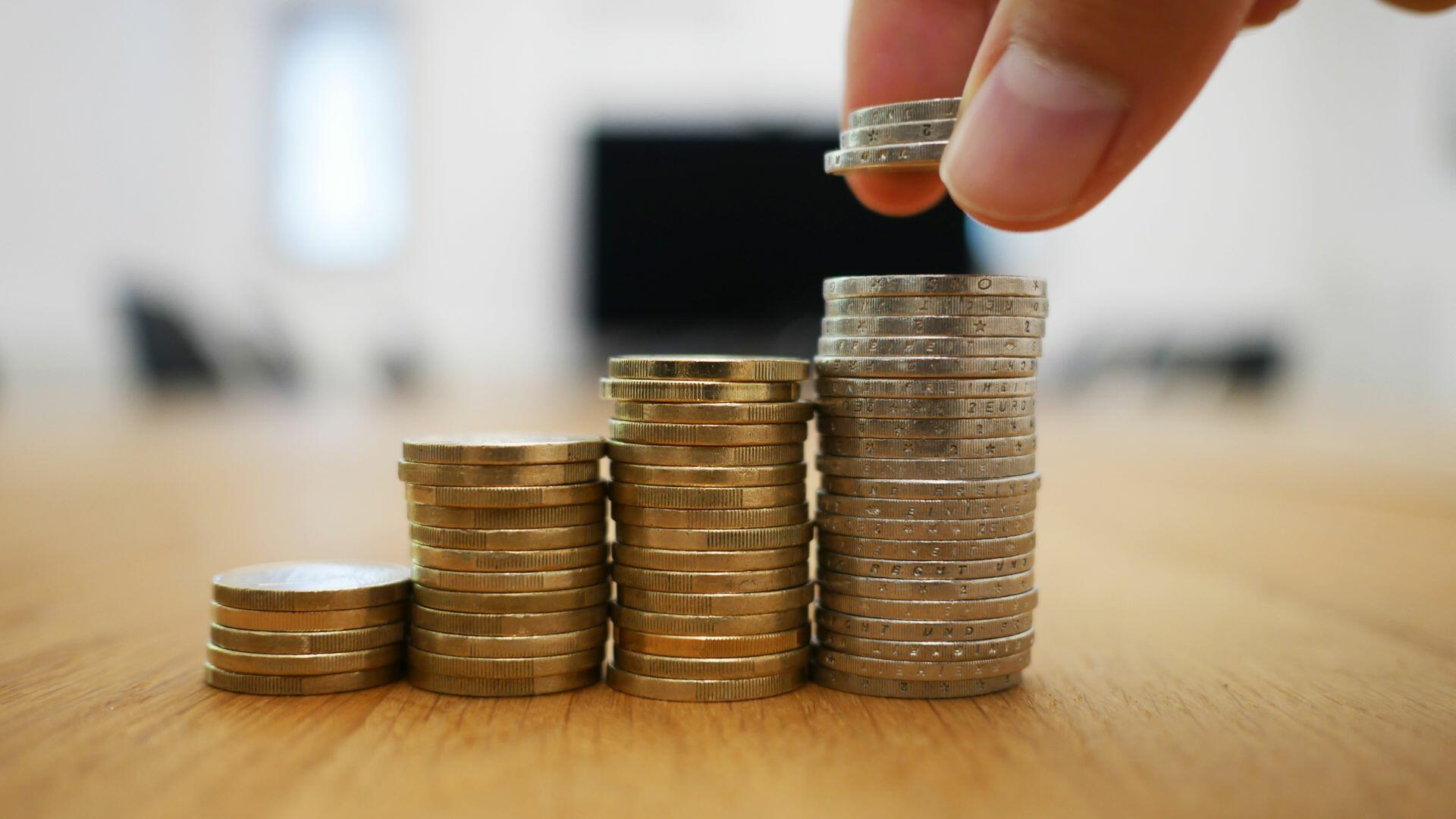 Mano colocando monedas sobre pilas crecientes de monedas de euro, lo que representa el crecimiento de la inversión.