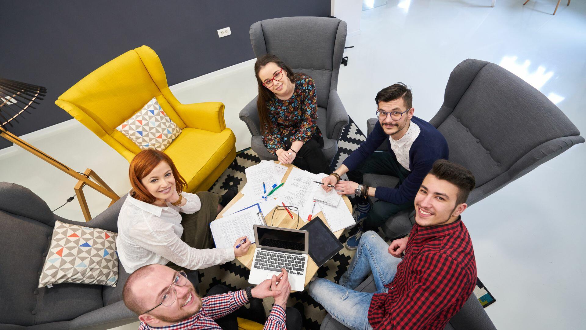 Cuatro jóvenes profesionales colaboran alrededor de una mesa, revisando documentos y utilizando un ordenador portátil.