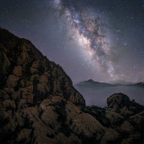 Vista nocturna de la Vía Láctea sobre un paisaje montañoso, con rocas en primer plano y un cielo estrellado que muestra colores vibrantes y detalles de la galaxia. Ideal para amantes de la astronomía y la fotografía de paisajes.