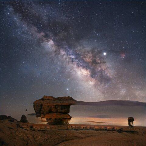 Paisaje nocturno con la Vía Láctea brillando en el cielo estrellado, junto a formaciones rocosas en primer plano, creando un contraste espectacular en un entorno desértico.