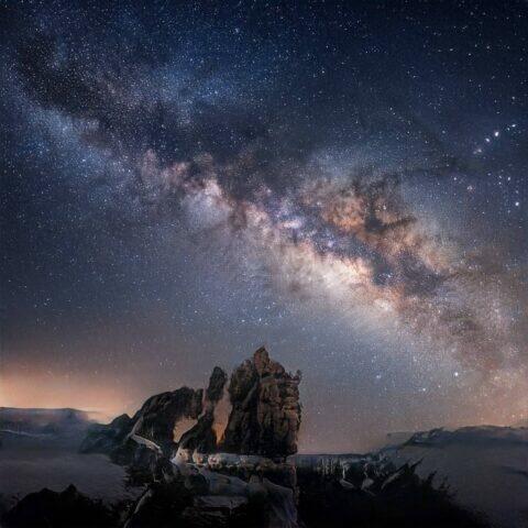 Cielo estrellado con la Vía Láctea sobre formaciones rocosas en un paisaje montañoso, capturando la belleza del universo en una noche despejada.