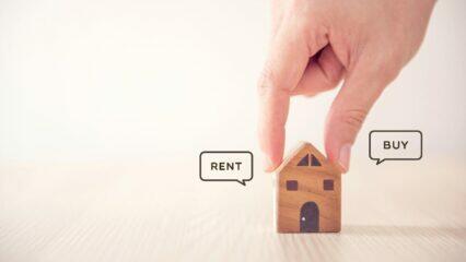 Mano colocando una maqueta de casa entre los bocadillos de "Alquilar" y "Comprar". Comprar vs. alquilar una vivienda.