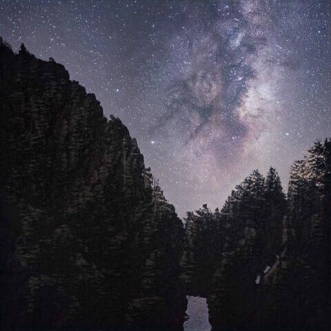 Vista nocturna de un cañón rodeado de árboles, con la Vía Láctea brillando en el cielo estrellado. La imagen muestra formaciones rocosas imponentes y un reflejo en el agua del cañón. Ideal para amantes de la naturaleza y la astronomía.