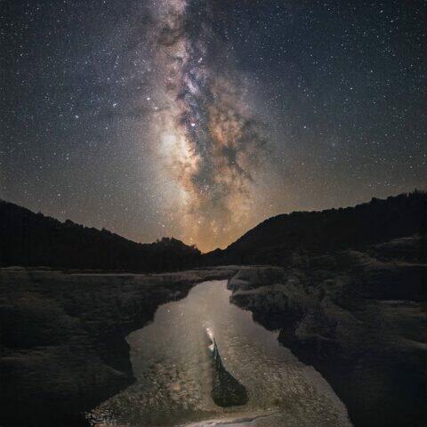 Vista nocturna del cielo estrellado con la Vía Láctea, reflejada en un río tranquilo rodeado de montañas. La imagen captura la belleza del paisaje natural y la inmensidad del universo.