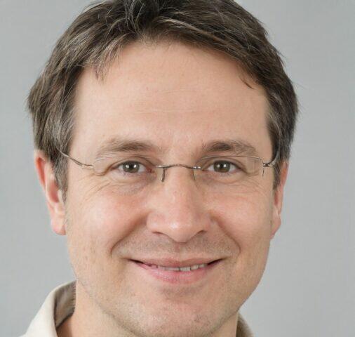 Hombre sonriente con gafas, cabello corto y canoso, fondo gris claro.