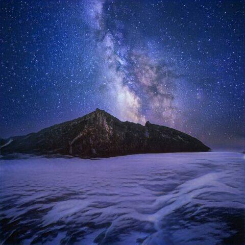 Paisaje nocturno con la Vía Láctea brillando sobre una montaña, rodeada de nubes y un lago, creando un ambiente mágico y sereno.